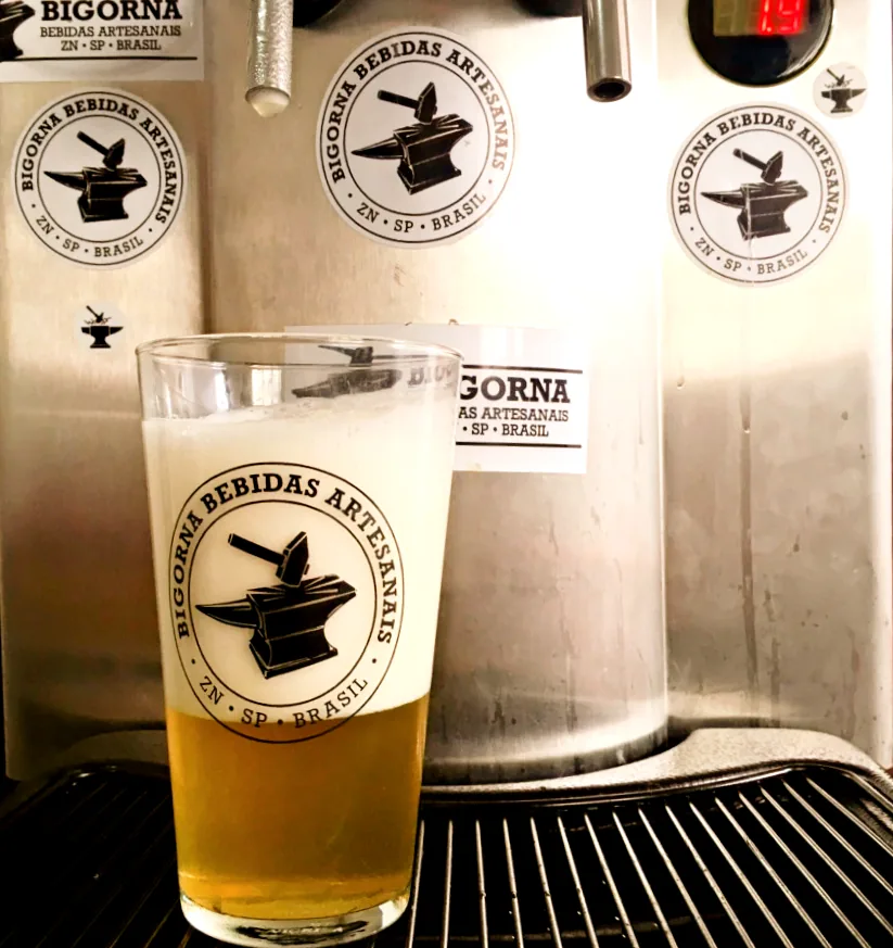 Copo com o carimbo da Bigorna cheio de chopp claro, sob a torneira da chopeira